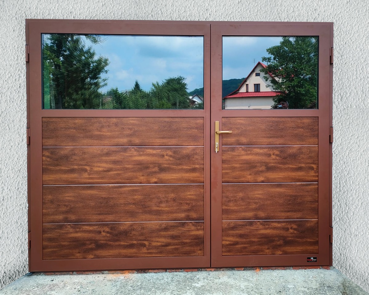 Dwuskrzydłowa brama garażowa z brązową ramą i drewnopodobnym panelem, górna część przeszklona, odbijająca krajobraz z domem i drzewami.