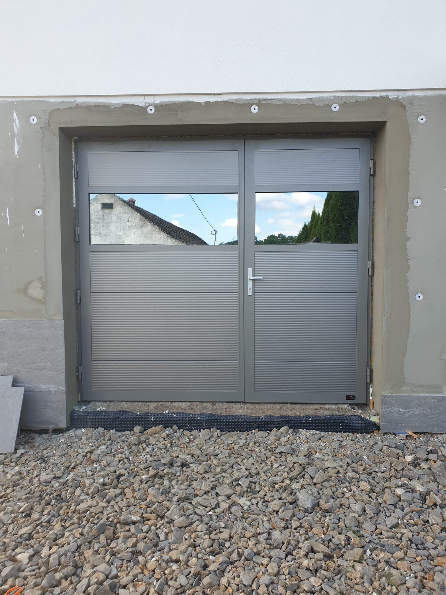 Szara, dwuskrzydłowa brama z poziomymi przetłoczeniami i okienkami, odbijającymi niebo i fragment budynku, zamontowana w surowym otworze ściany z widocznymi elementami montażowymi i kamienistym...