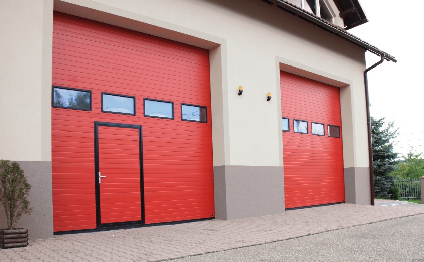 Dwa czerwone segmentowe zamknięcia w budynku z beżową elewacją, jedno z drzwiami wejściowymi i oknami, drugie z oknami, oświetlenie zewnętrzne.