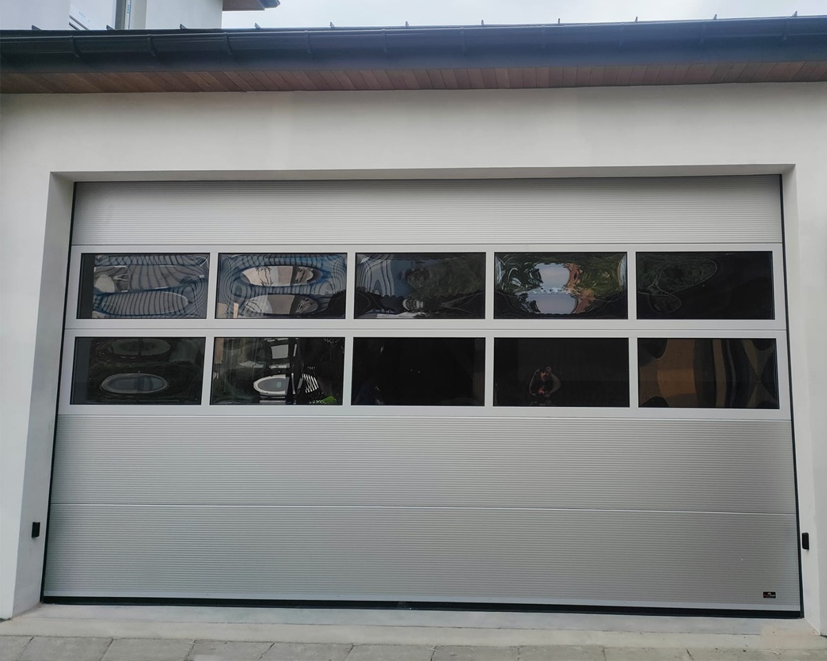 Srebrna segmentowa brama z poziomymi panelami i rzędem okienek w górnej części, odbijających otoczenie.