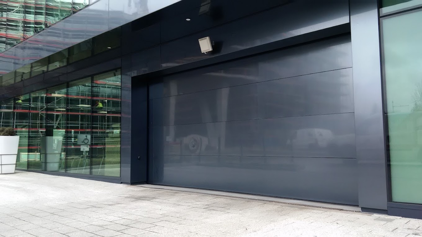 Zamknięta, segmentowa brama przemysłowa w kolorze antracytowym, zamontowana w nowoczesnym budynku biurowym z przeszkloną fasadą, widoczne odbicia w szybach.