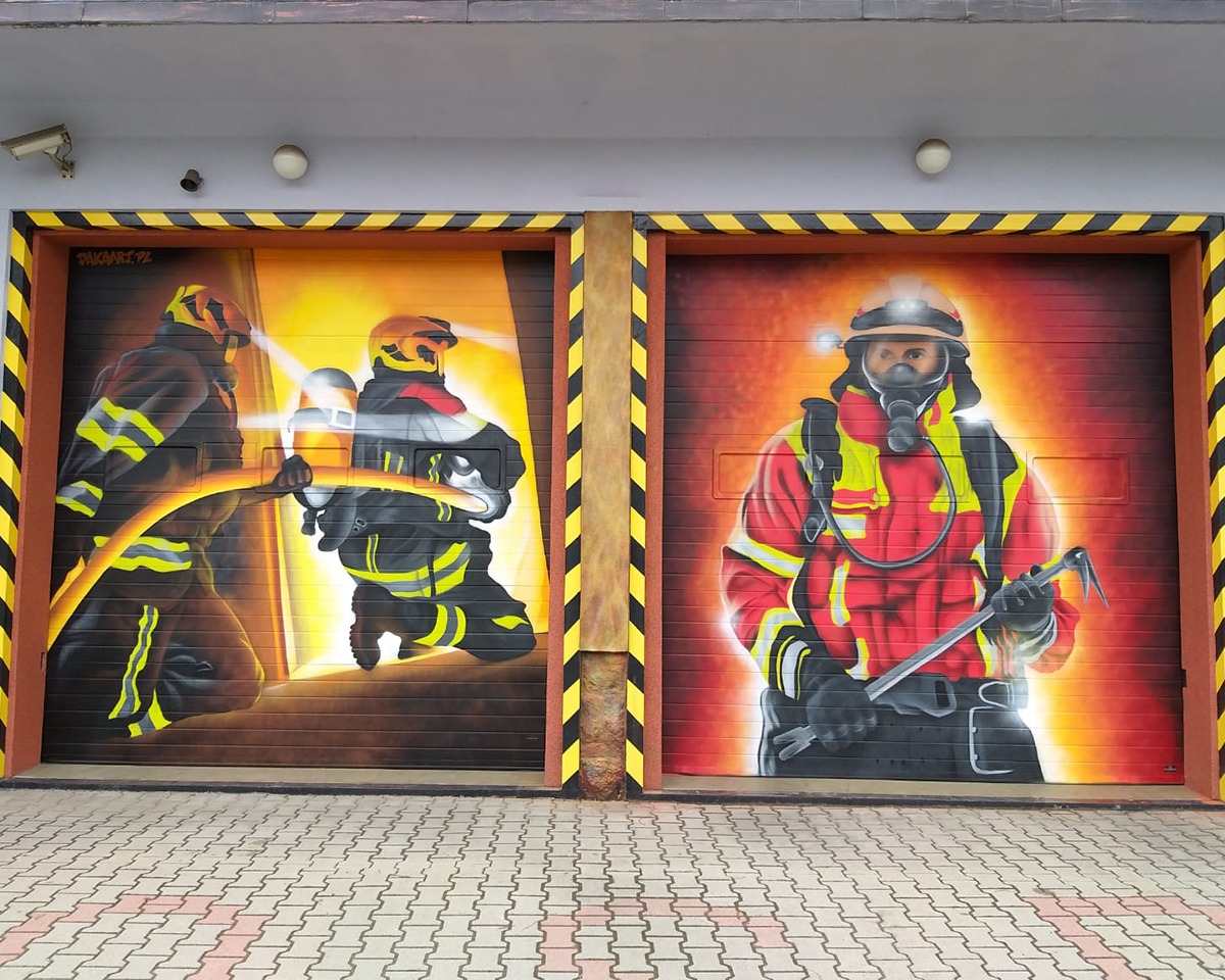 Dwie pomalowane segmentowe konstrukcje z wizerunkami strażaków w akcji, otoczone czarno-żółtymi pasami ostrzegawczymi. Po lewej stronie dwóch strażaków z latarkami, po prawej strażak w masce i...