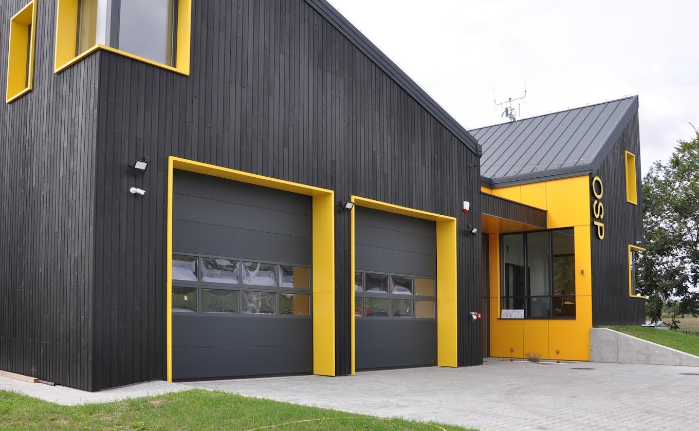 Nowoczesny budynek OSP z dwoma szarymi segmentowymi bramami garażowymi obramowanymi żółtymi panelami, czarne drewniane deski elewacyjne, szara kostka brukowa na podjeździe.