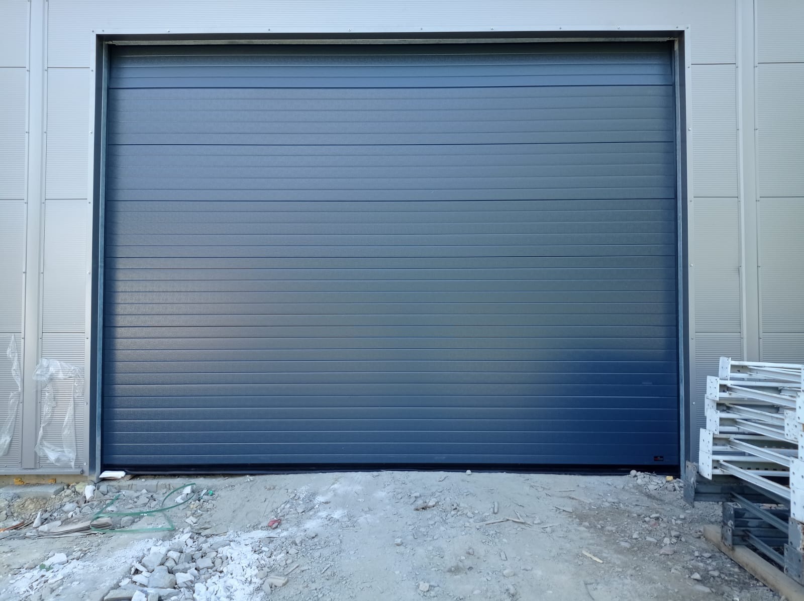 Szara segmentowa konstrukcja zamknięta, zamontowana w hali przemysłowej, widoczne elementy konstrukcyjne budynku i gruz budowlany.