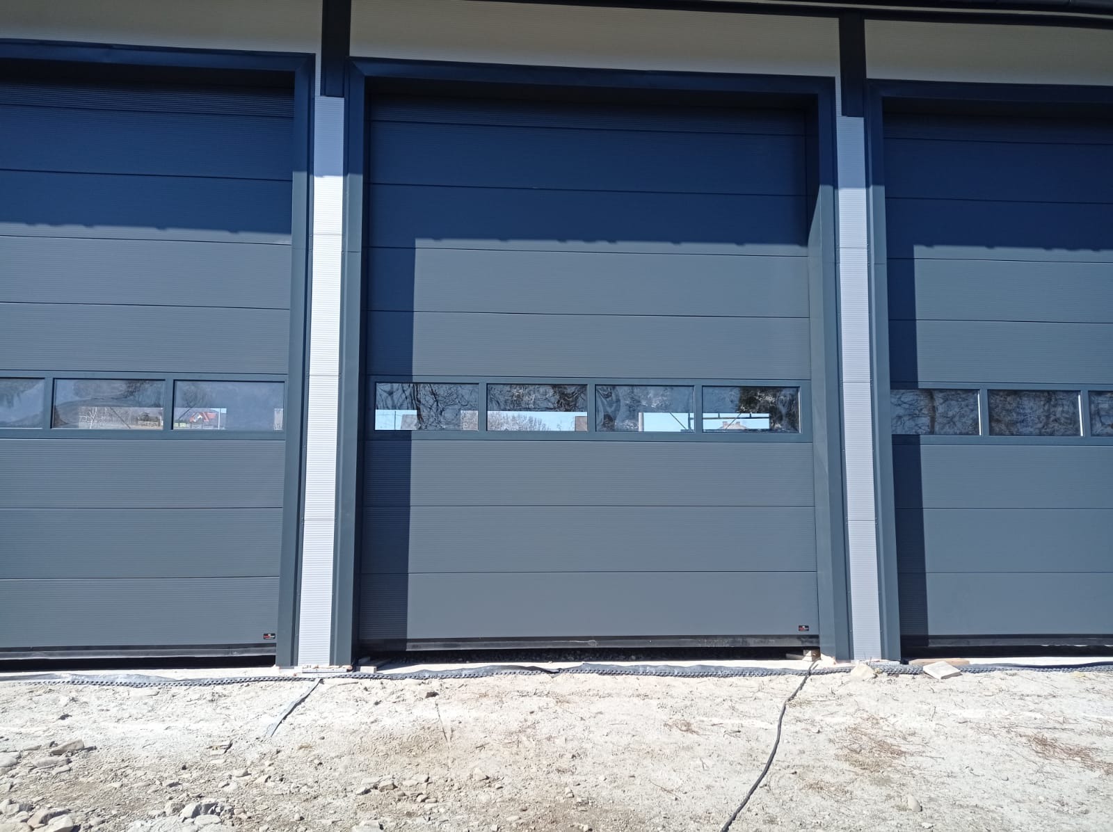 Szare segmentowe drzwi garażowe z oknami, częściowo otwarte, widoczne trzy stanowiska w hali.