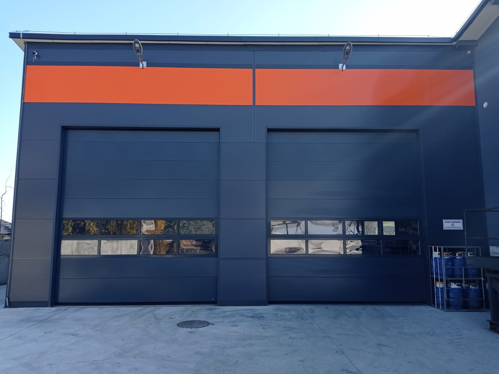 Dwie zamknięte, segmentowe konstrukcje w kolorze grafitowym z poziomym oknem, zamontowane w hali przemysłowej z pomarańczowym pasem na elewacji.