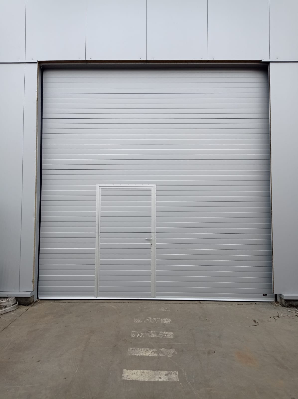 Szara brama przemysłowa z wbudowanymi drzwiami dla pieszych, widoczna z zewnątrz budynku w Raciechowicach.