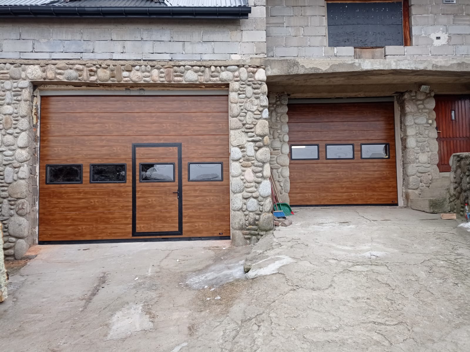 Dwie brązowe, segmentowe konstrukcje z oknami, jedna z drzwiami, zamontowane w kamiennej elewacji budynku gospodarczego.