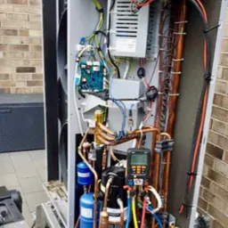 Otwarta obudowa pompy ciepła podczas serwisowania, widoczne komponenty wewnętrzne, miedziane rurki, elektroniczny manometr podłączony do układu chłodniczego, tło z cegły.