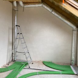 Dom-Tech Jakub Urbański - Instalacja przewodów wentylacyjnych i zielonych rur ogrzewania podłogowego w surowym wnętrzu poddasza, widoczna drabina aluminiowa.
