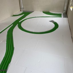 Dom-Tech Jakub Urbański - Zielone rury instalacji grzewczej ułożone w rowkach styropianowej izolacji podłogowej, widoczne elementy mocujące i aluminiowe rury wentylacyjne w rogu pomieszczenia.