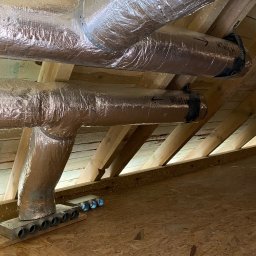 Dom-Tech Jakub Urbański - Izolowane kanały wentylacyjne pod drewnianym stropem poddasza, z widocznymi strzałkami kierunku przepływu powietrza i skrzynką rozdzielczą z okrągłymi wylotami, akumulator Bosch 18V na podłodze.