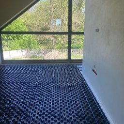 Dom-Tech Jakub Urbański - Niedokończona instalacja ogrzewania podłogowego z niebieskimi rurami na czarnej macie, widok z góry na tle dużego okna z widokiem na zieleń.