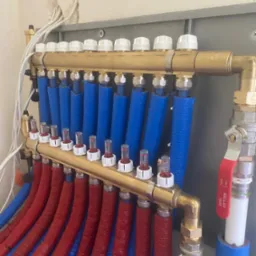 Rozdzielacz ogrzewania podłogowego z rurami w izolacji, część rur w kolorze czerwonym a część w kolorze niebieskim, zamontowany na ścianie z widocznymi przewodami elektrycznymi.