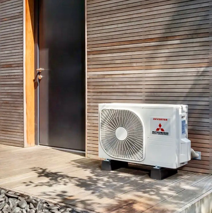 Biała jednostka zewnętrzna klimatyzacji Mitsubishi Inverter zamontowana na czarnych podstawkach przy drewnianej elewacji domu z czarnymi drzwiami, widoczne cienie drzew.