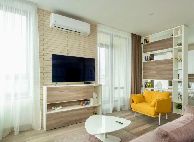 Biała jednostka klimatyzacyjna zamontowana na ścianie z cegły w nowoczesnym salonie z telewizorem, żółtą sofą i białymi zasłonami.