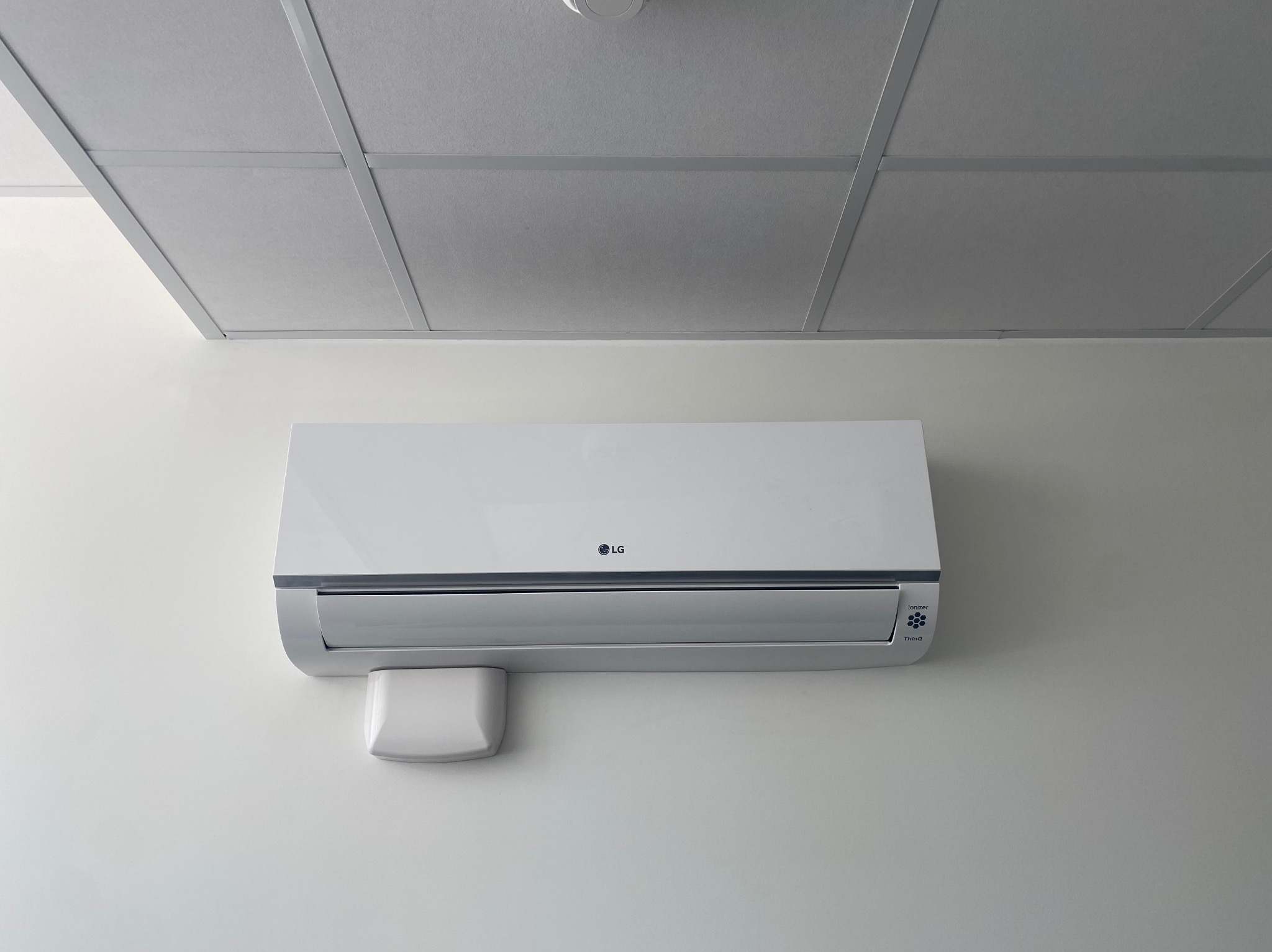 Biały klimatyzator LG zamontowany na jasnej ścianie pod sufitem kasetonowym. Widoczny czujnik na ścianie obok urządzenia.