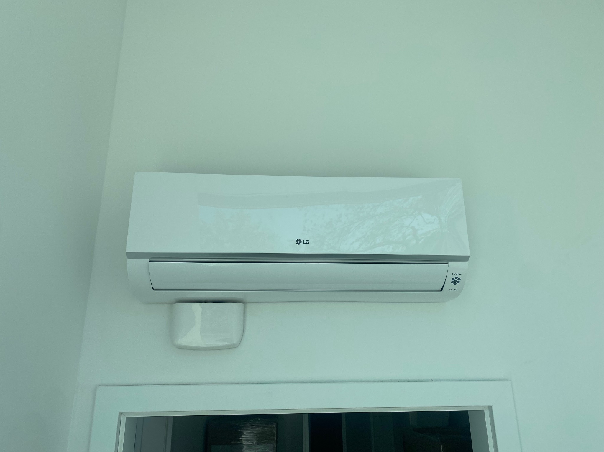 Biały klimatyzator LG zamontowany na ścianie nad ościeżnicą drzwi, widoczny zbiornik na skropliny po lewej stronie urządzenia.