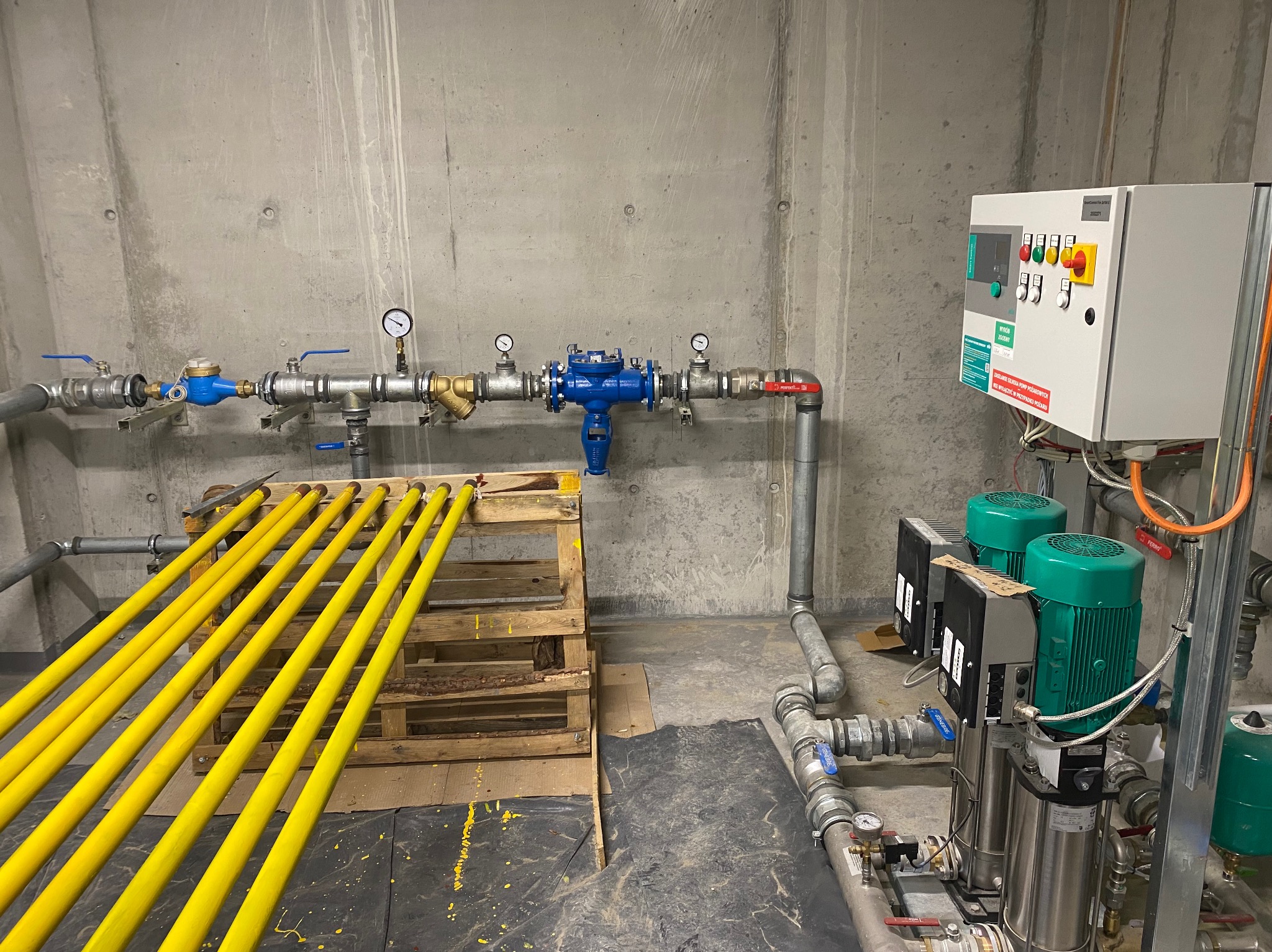 Instalacja wodociągowa z licznymi zaworami, manometrami i pompami, widoczne żółte rury na palecie, szara betonowa ściana w tle.