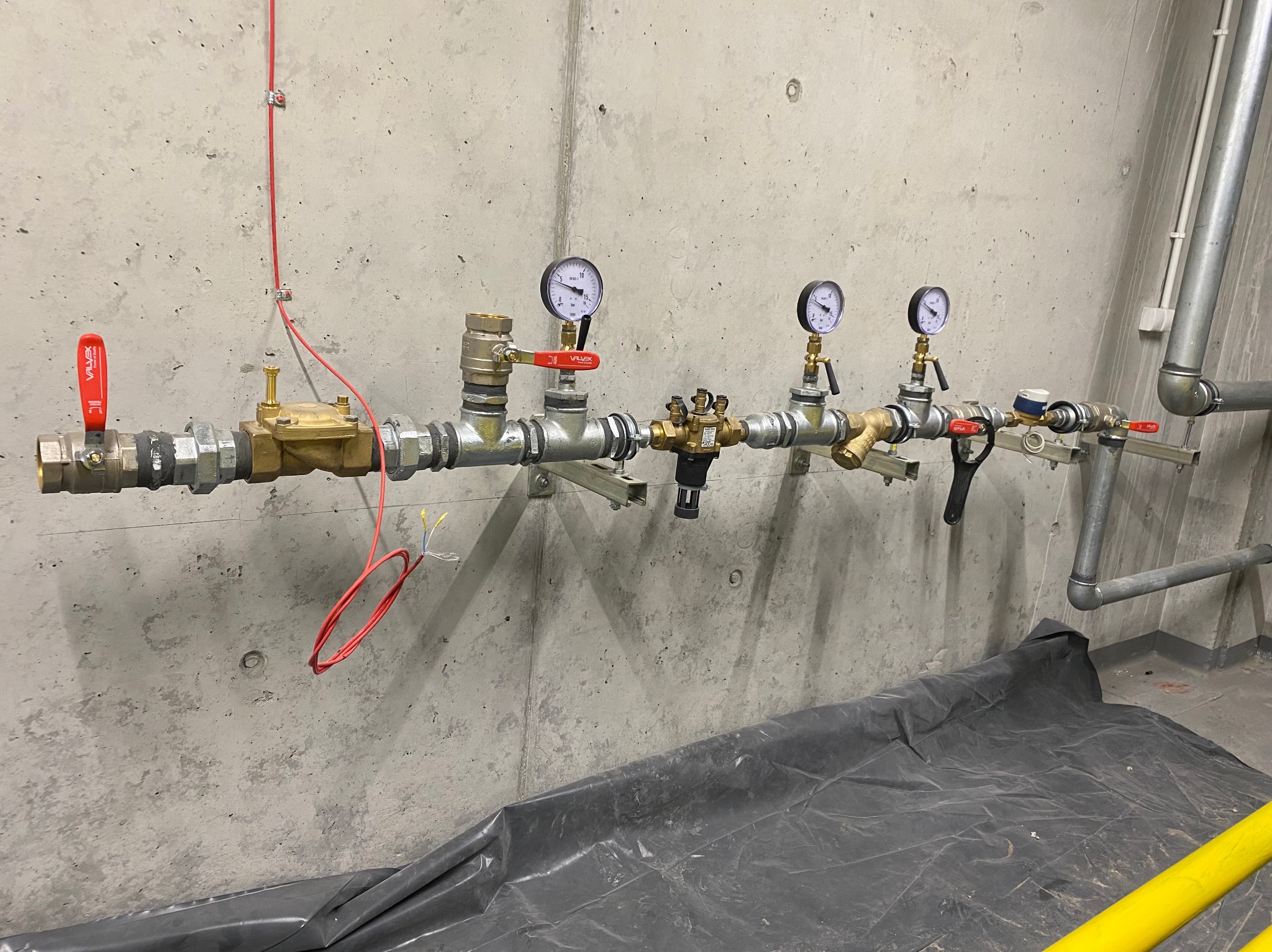 Instalacja wodno-kanalizacyjna z manometrami na szarym, betonowym tle w Częstochowie, zabezpieczona czarną folią. Widoczne zawory i rury.