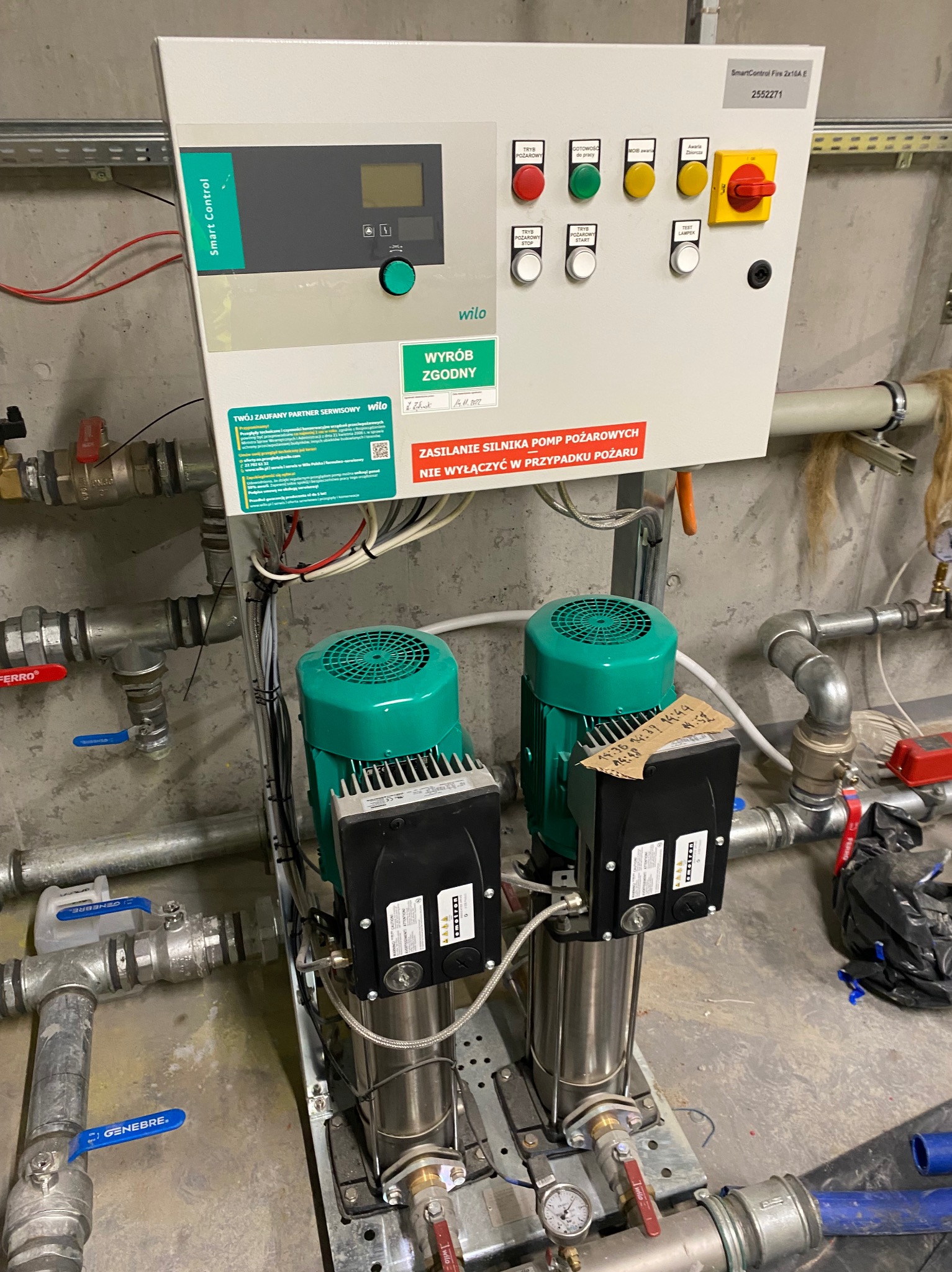 Dwie pionowe pompy Wilo z zielonymi silnikami, podłączone do rur, z panelem sterowania SmartControl Fire 2x16A E z przyciskami i naklejkami ostrzegawczymi, zainstalowane w pomieszczeniu technicznym.