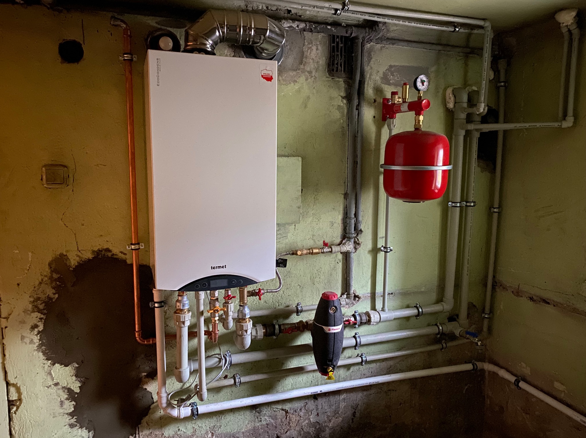 Kocioł gazowy kondensacyjny marki Termet zamontowany na pomalowanej na zielono ścianie z widocznymi śladami wilgoci, z licznymi rurami miedzianymi i plastikowymi, oraz czerwonym zbiornikiem...