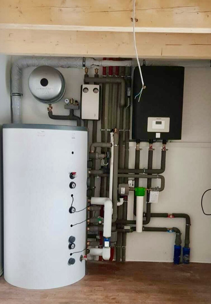 Biały akumulacyjny zbiornik ciepłej wody użytkowej z podłączonymi rurami i osprzętem w kotłowni, widoczne elementy izolacji termicznej na rurach.