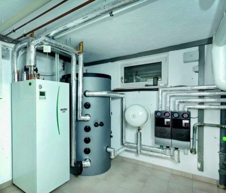 Kotłownia z nowoczesnym piecem, zasobnikiem wody i systemem rur w izolacji, widok z boku, w tle małe okno piwniczne.