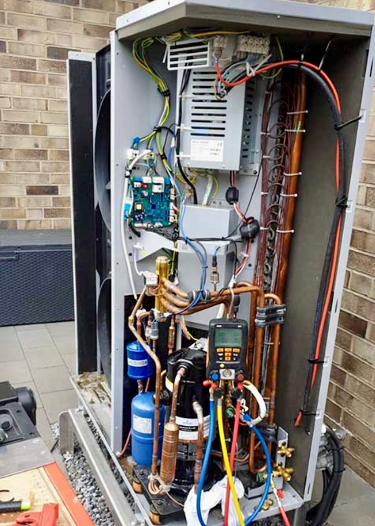 Otwarta obudowa pompy ciepła podczas serwisowania, widoczne komponenty wewnętrzne, miedziane rurki, elektroniczny manometr podłączony do układu chłodniczego, tło z cegły.