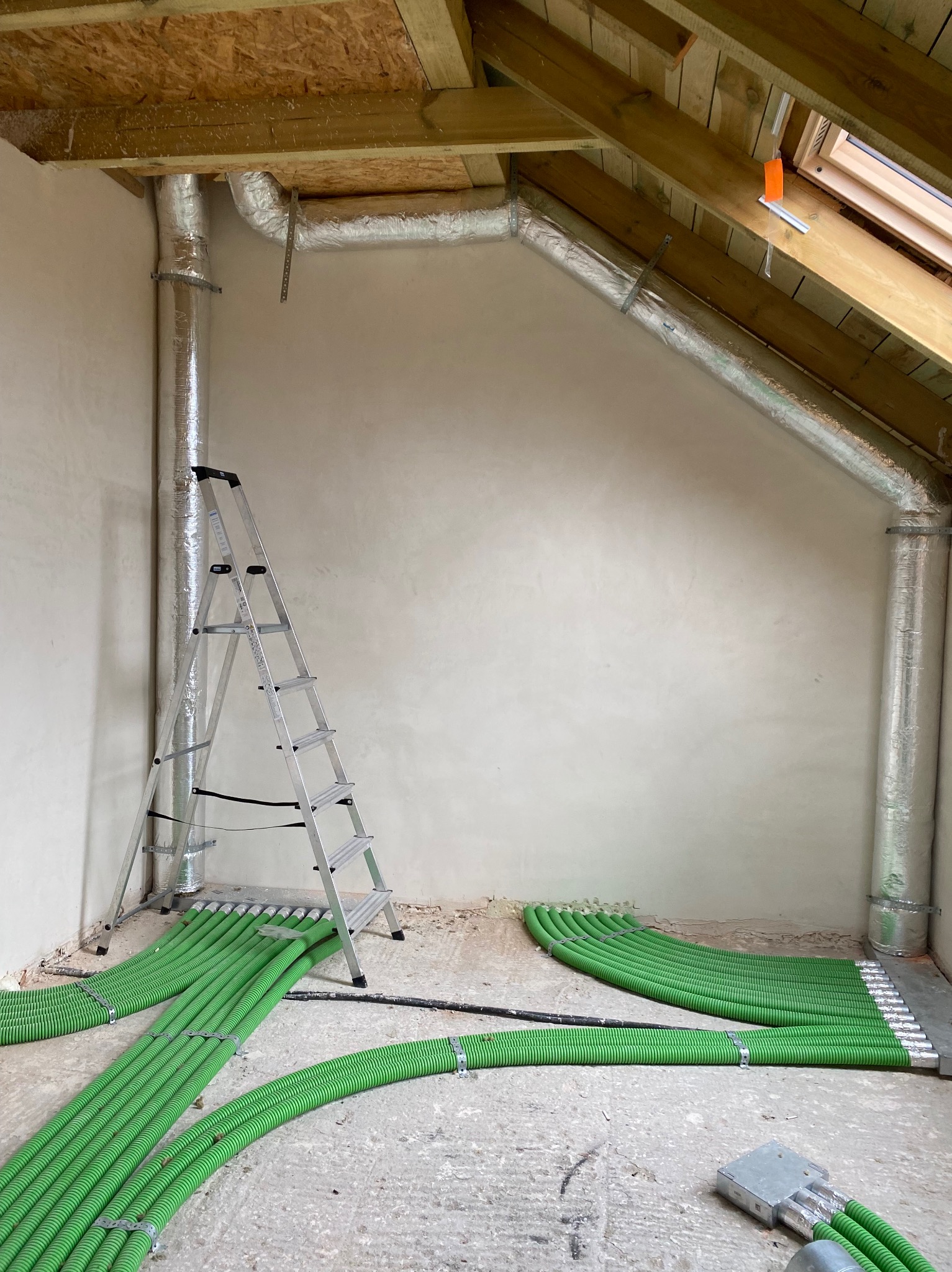Instalacja przewodów wentylacyjnych i zielonych rur ogrzewania podłogowego w surowym wnętrzu poddasza, widoczna drabina aluminiowa.