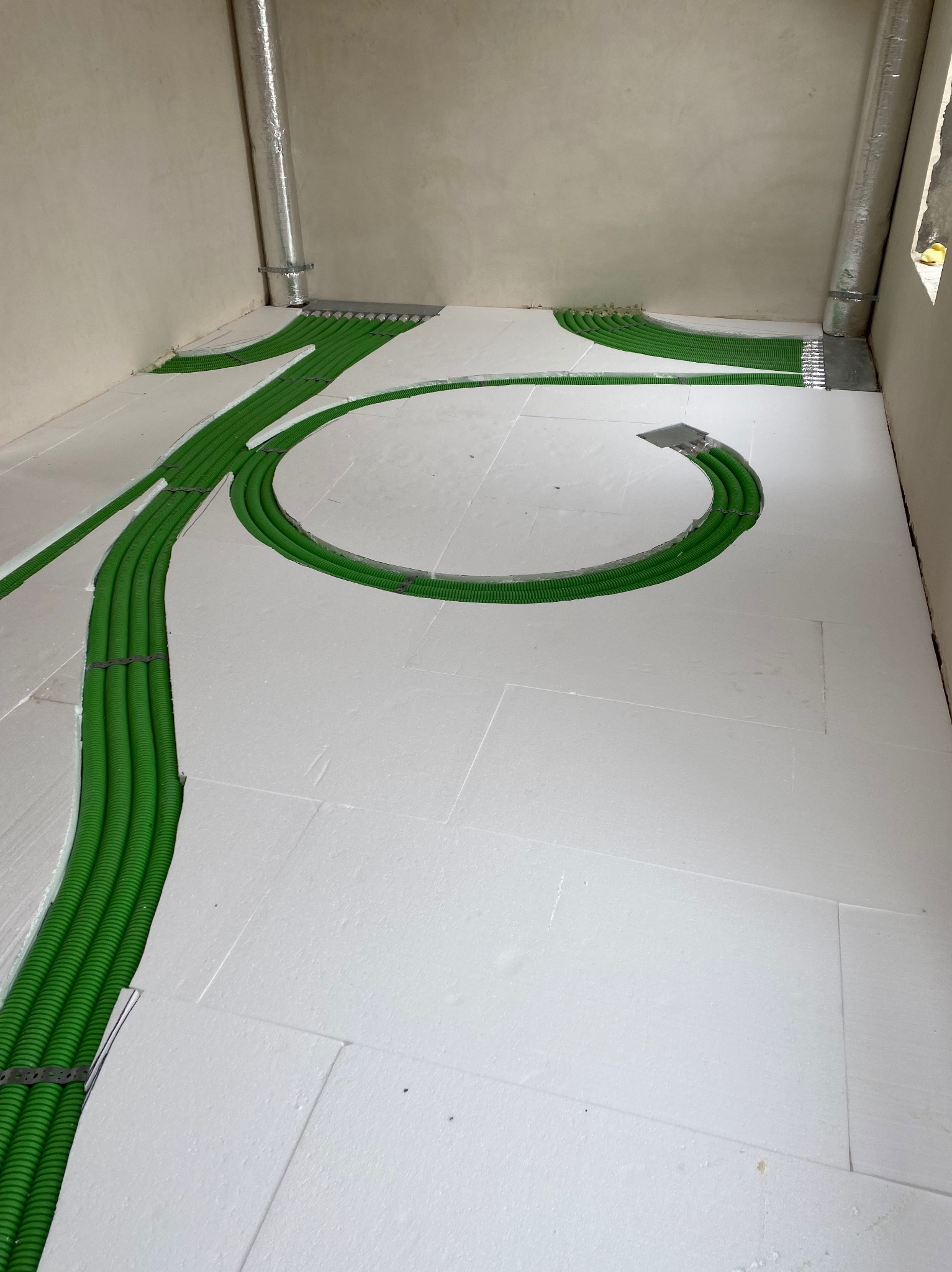 Zielone rury instalacji grzewczej ułożone w rowkach styropianowej izolacji podłogowej, widoczne elementy mocujące i aluminiowe rury wentylacyjne w rogu pomieszczenia.