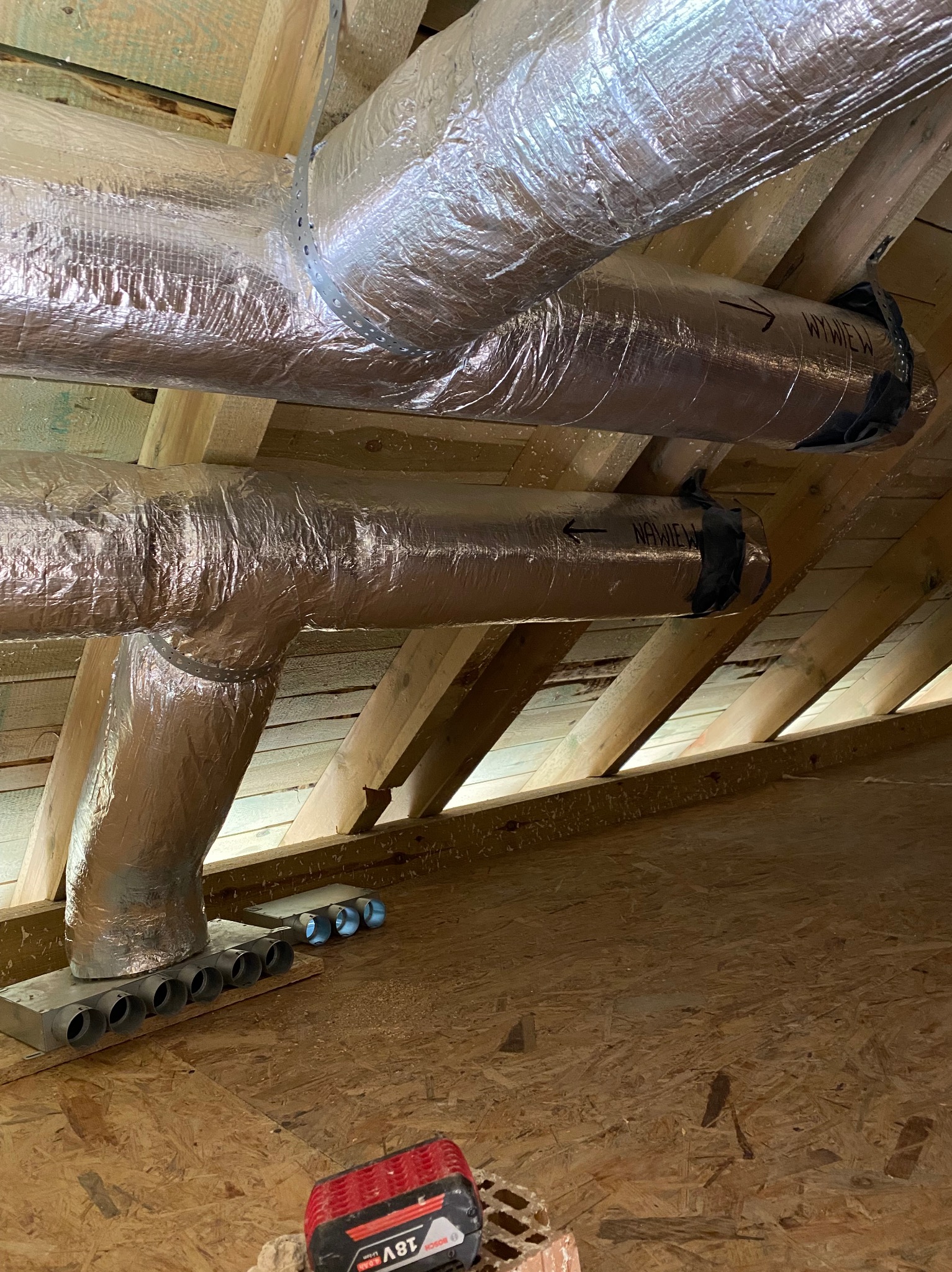 Izolowane kanały wentylacyjne pod drewnianym stropem poddasza, z widocznymi strzałkami kierunku przepływu powietrza i skrzynką rozdzielczą z okrągłymi wylotami, akumulator Bosch 18V na podłodze.