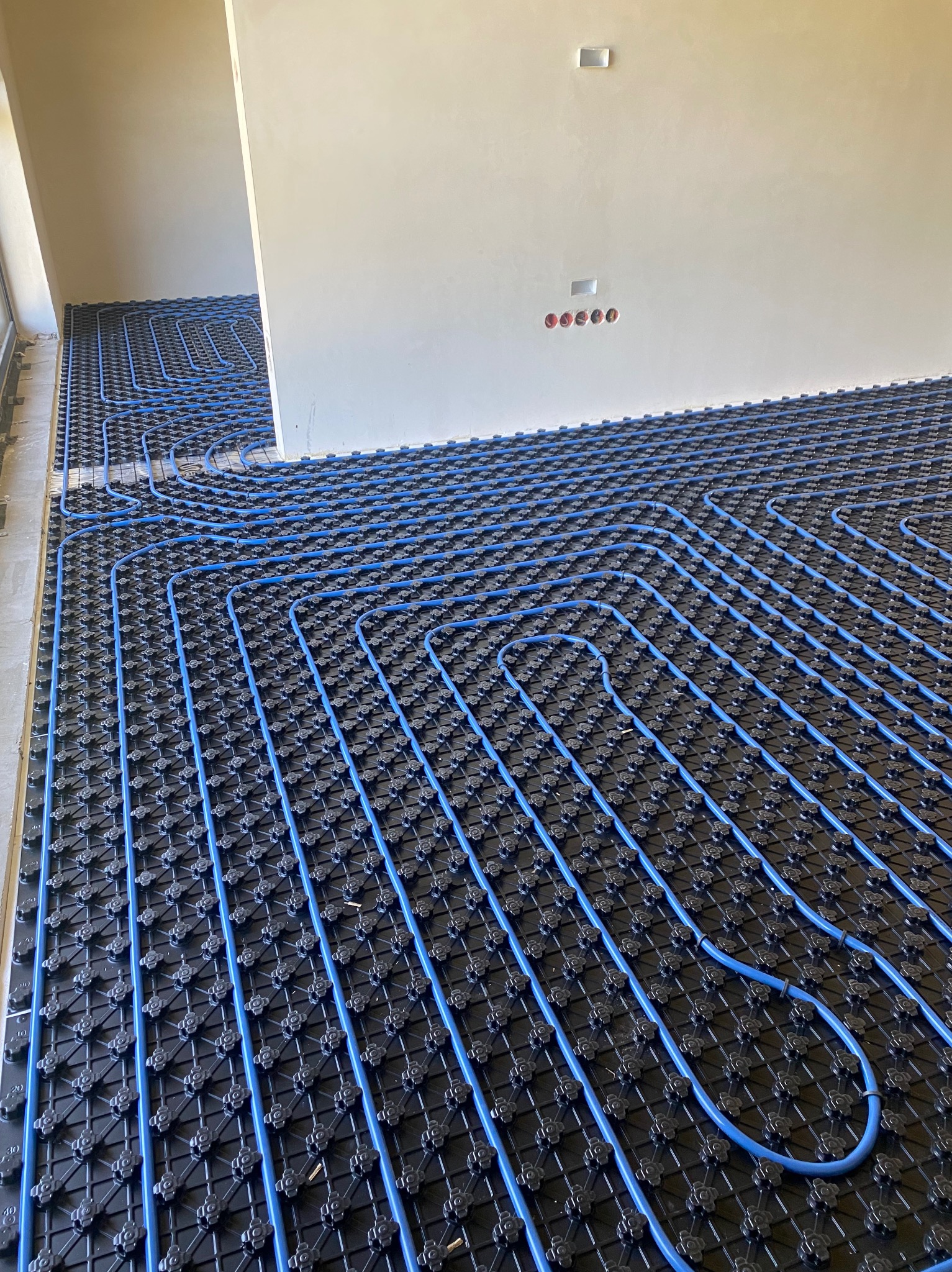Niebieskie rury ogrzewania podłogowego ułożone na czarnej macie montażowej, widok pod kątem w surowym stanie wykończenia wnętrza.