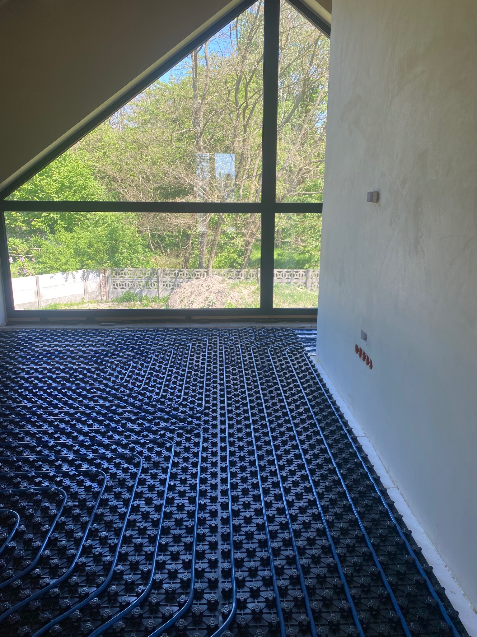 Niedokończona instalacja ogrzewania podłogowego z niebieskimi rurami na czarnej macie, widok z góry na tle dużego okna z widokiem na zieleń.
