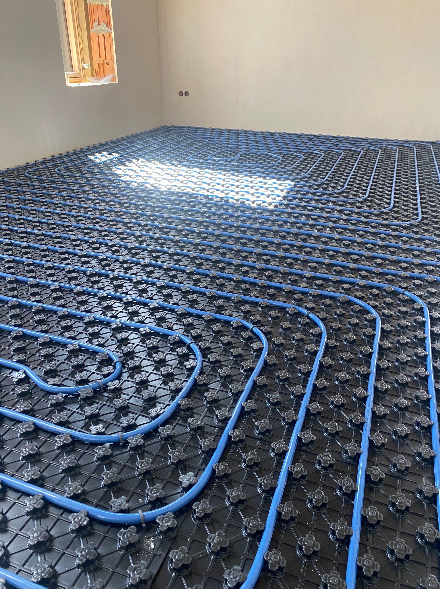 Niedokończona instalacja ogrzewania podłogowego z niebieskimi rurami PEX na czarnej macie izolacyjnej, widok z góry w surowym wnętrzu z oknem.