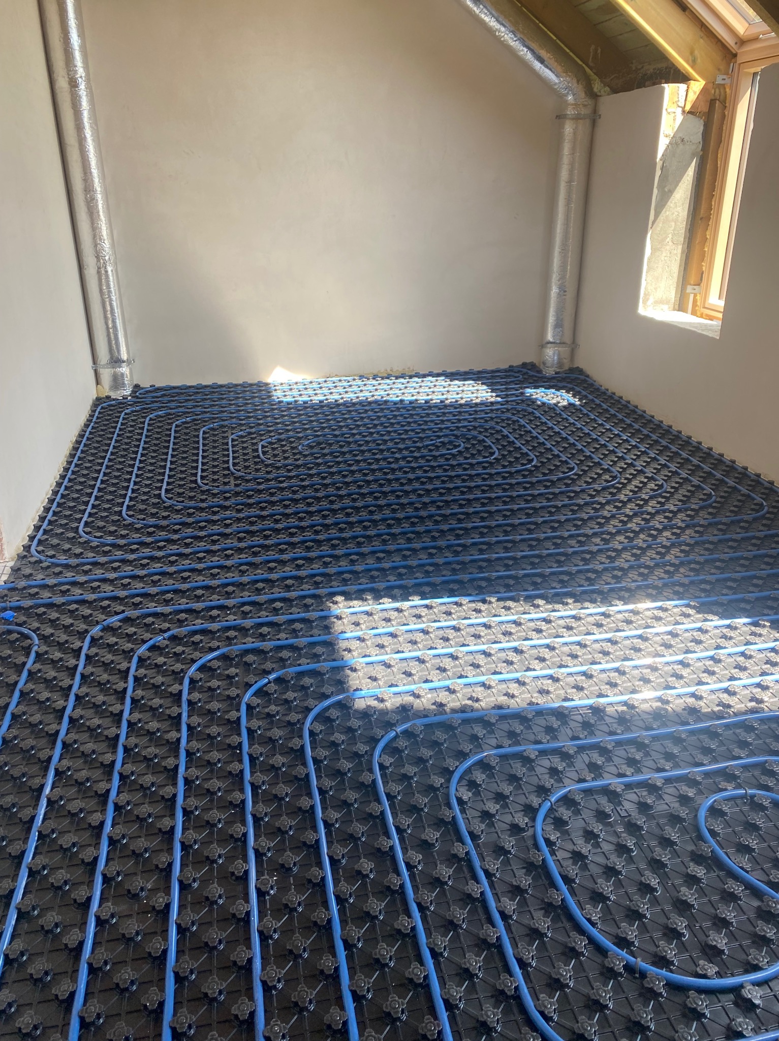 Niebieskie rurki ogrzewania podłogowego ułożone na czarnej macie montażowej w surowym pomieszczeniu z oknem dachowym i rurami wentylacyjnymi.