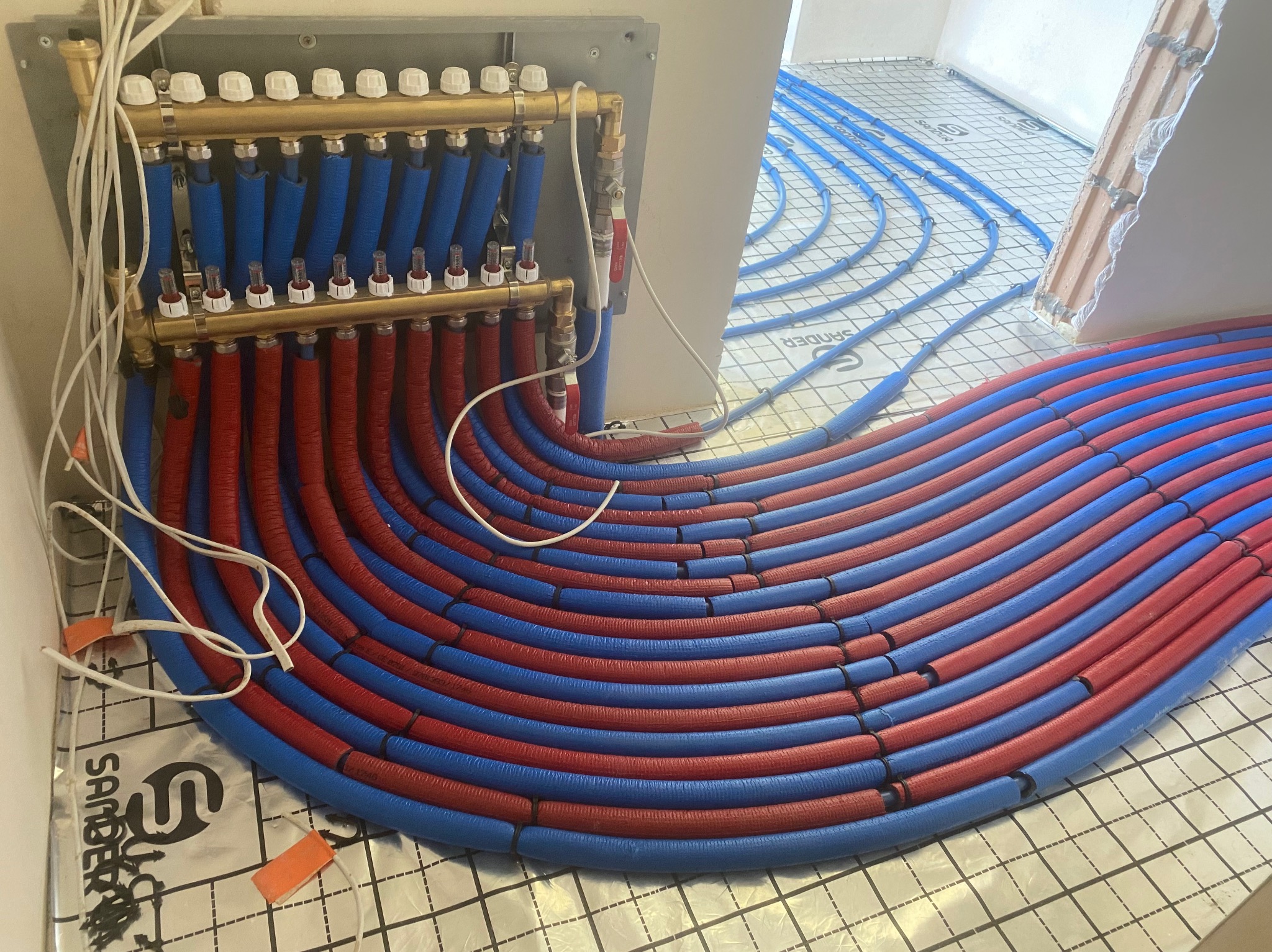 Rozłożone rury ogrzewania podłogowego w kolorach czerwonym i niebieskim, podłączone do rozdzielacza, ułożone na folii izolacyjnej z nadrukiem producenta.