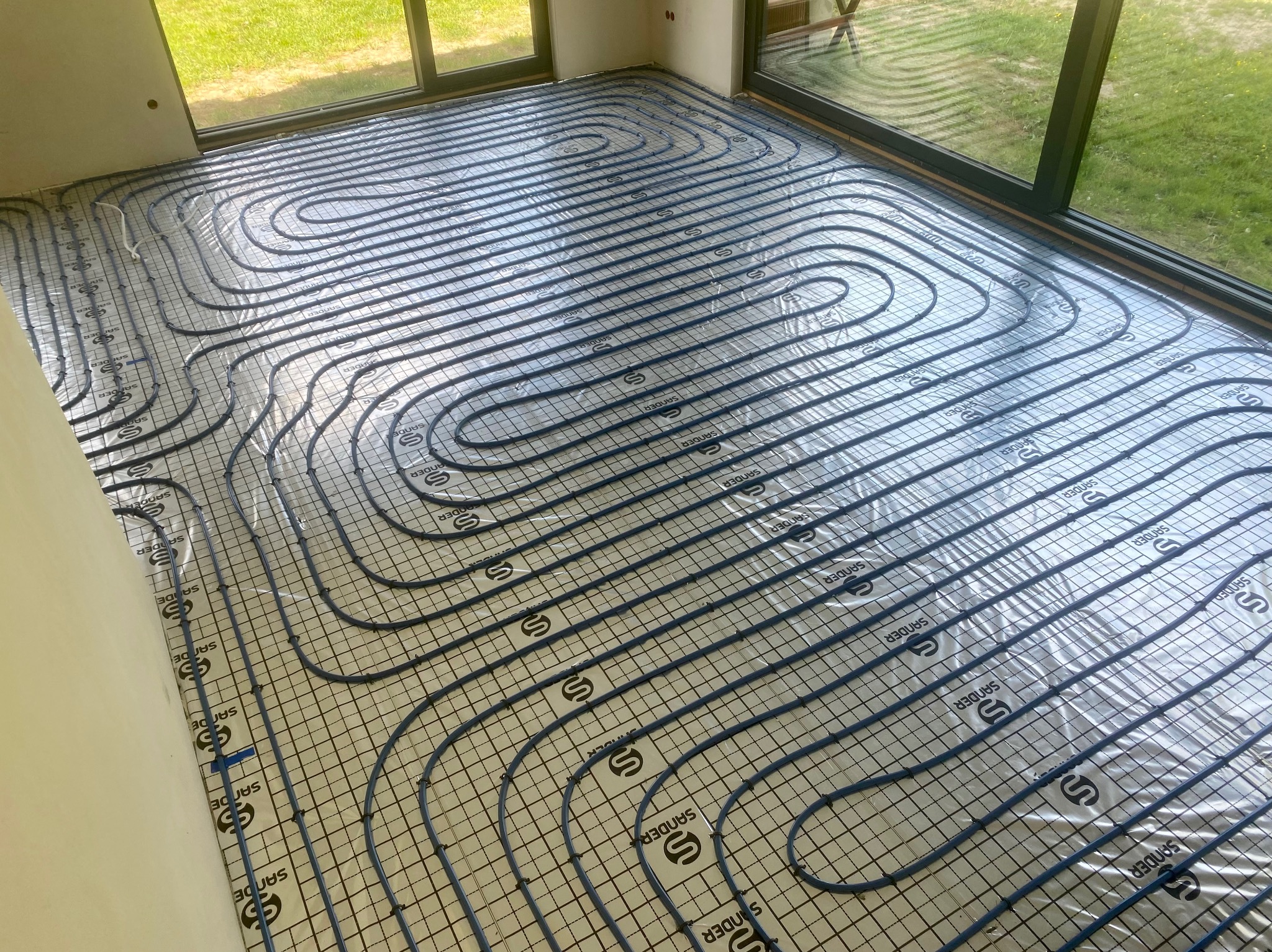 Rozłożona instalacja ogrzewania podłogowego na folii z nadrukowaną siatką i logo Sander, widok z góry na jasne wnętrze z dużymi oknami.
