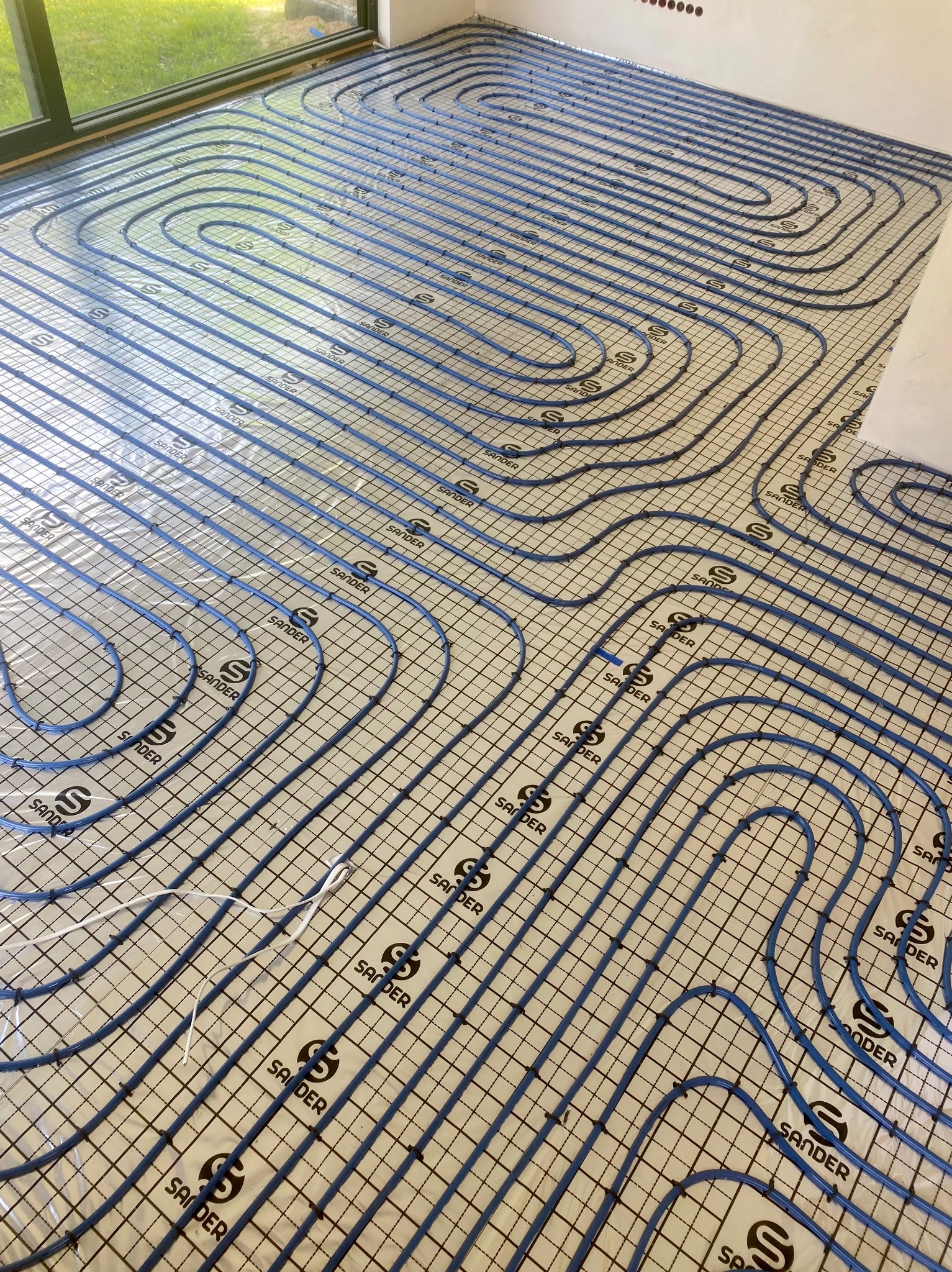 Montaż ogrzewania podłogowego: widok z góry na ułożone rury grzewcze w kolorze niebieskim na folii z nadrukowaną kratką i logo 'SANDER', gotowe do zalania wylewką.