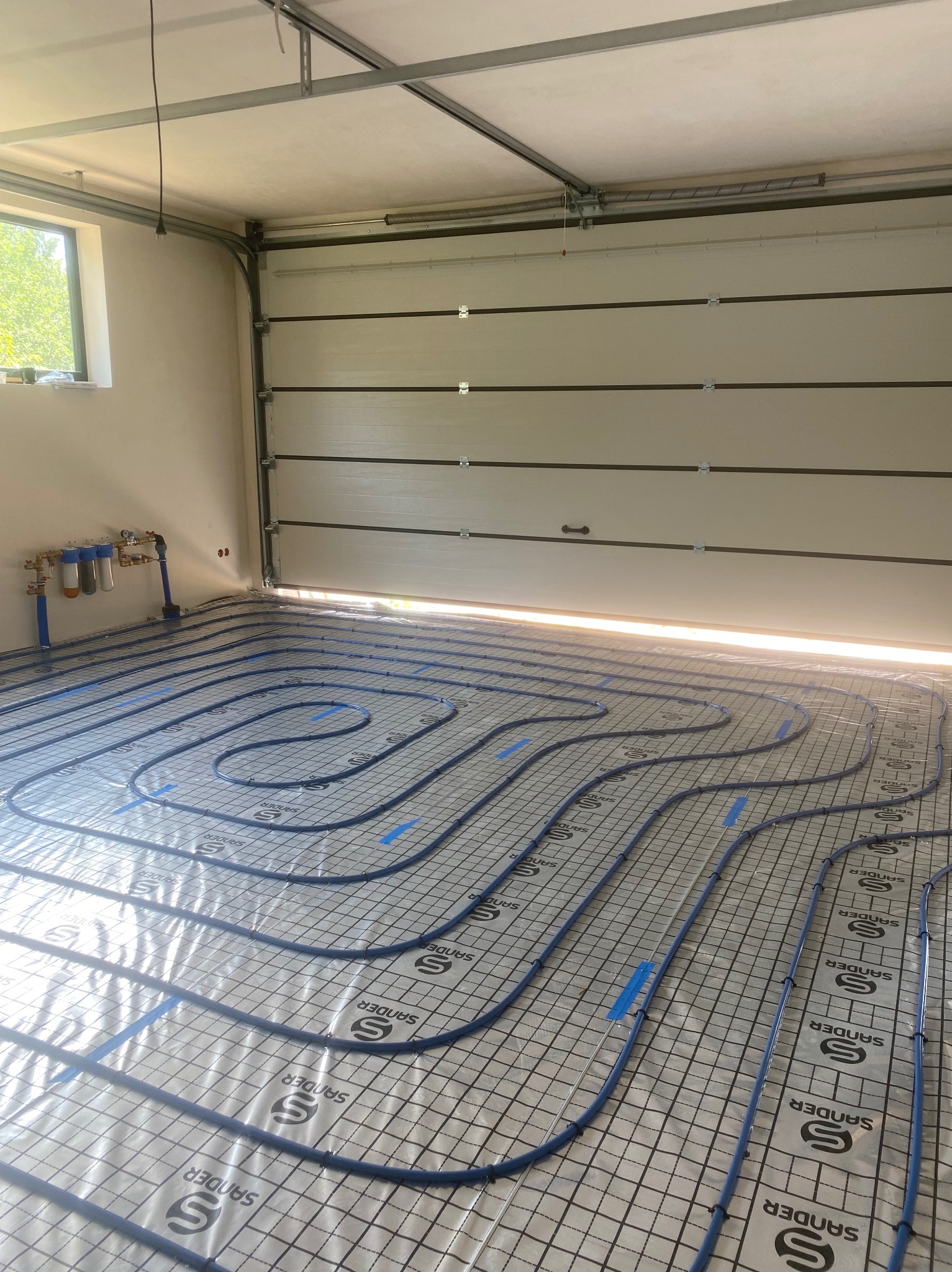 Instalacja ogrzewania podłogowego w garażu: niebieskie rury ułożone na folii z nadrukowaną siatką i logo Sander, widoczne przyłącza do instalacji wodnej na ścianie, brama garażowa w tle.