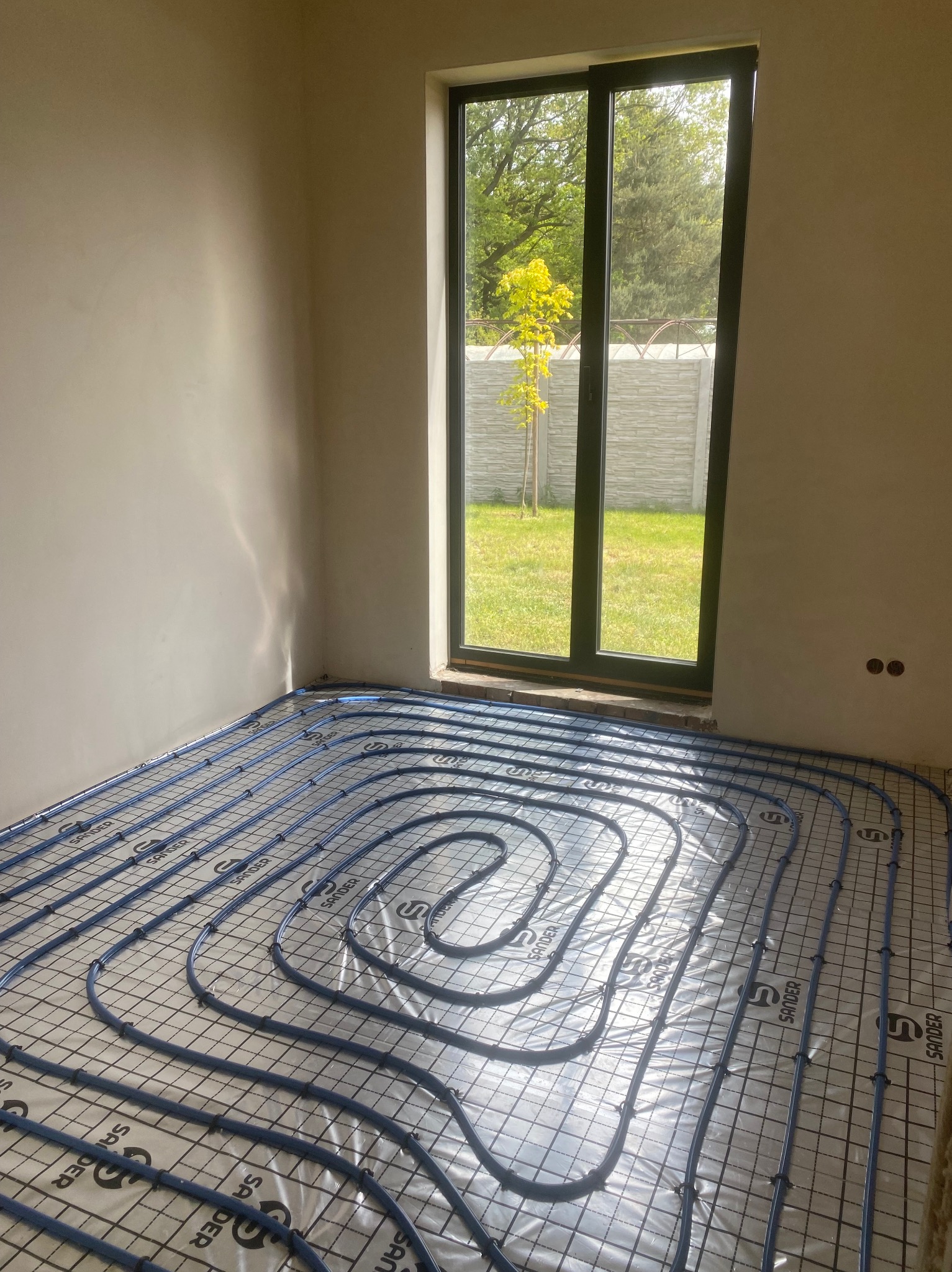 Niedokończona instalacja ogrzewania podłogowego z niebieskimi rurami na folii z nadrukiem Sander w jasnym, pustym pomieszczeniu z widokiem na ogród przez okno.