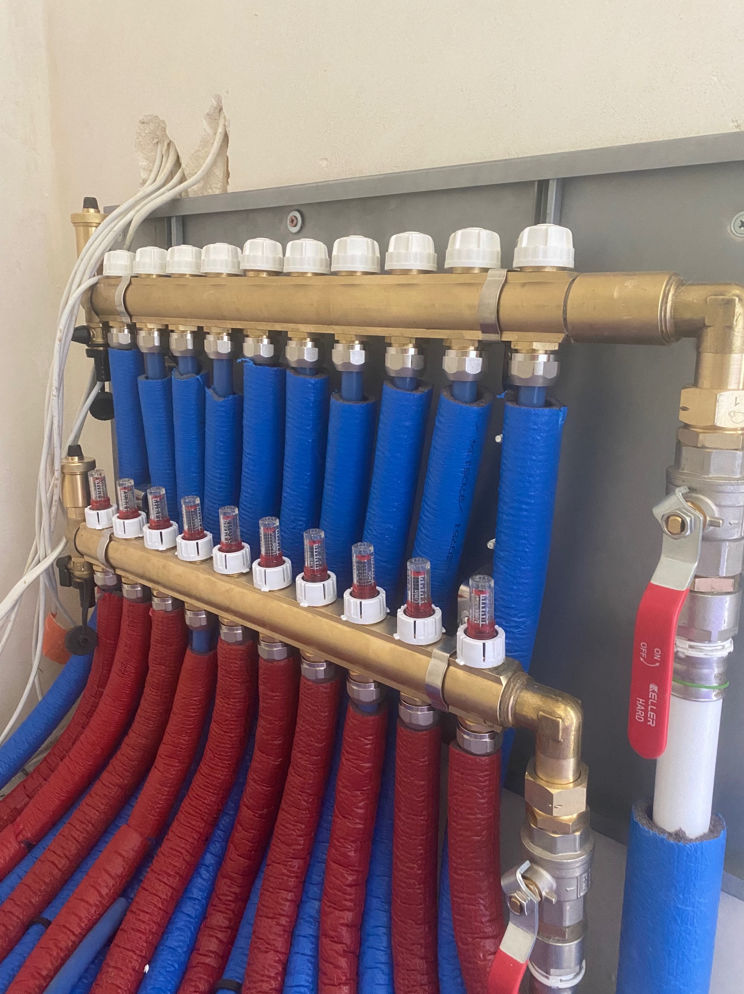 Rozdzielacz ogrzewania podłogowego z rurami w izolacji, część rur w kolorze czerwonym a część w kolorze niebieskim, zamontowany na ścianie z widocznymi przewodami elektrycznymi.