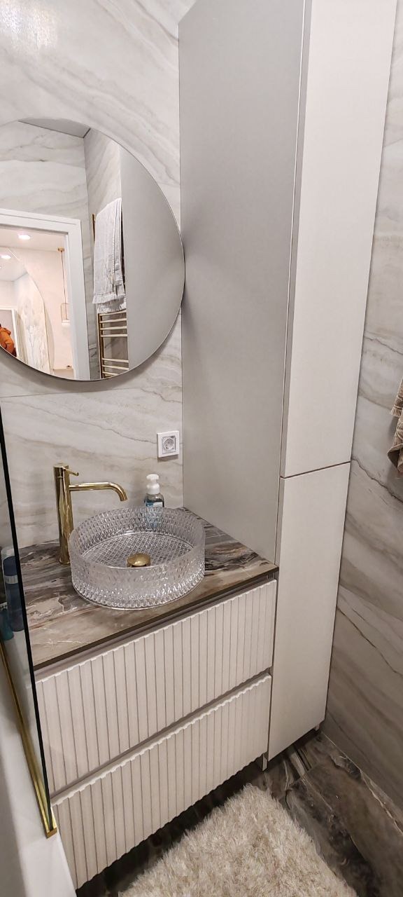 Nowoczesna łazienka z marmurowymi ścianami, okrągłym lustrem i szafką z umywalką z przezroczystego szkła oraz złotą armaturą.