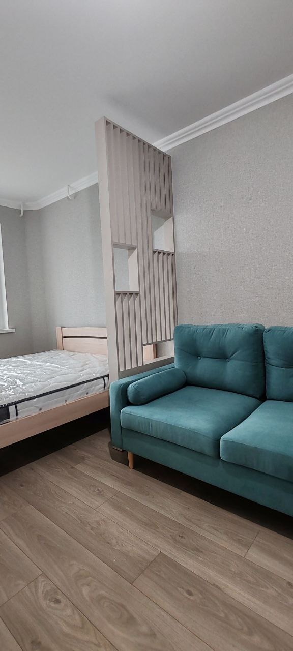 Turkusowa sofa i łóżko z materacem w folii, oddzielone ażurowym, drewnianym przepierzeniem w nowoczesnym wnętrzu.