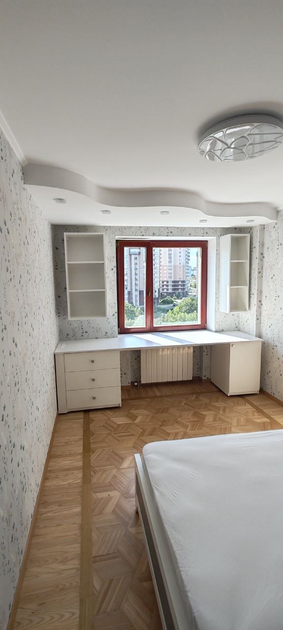 Białe biurko z szafkami i półkami zamontowane pod oknem w pokoju z wzorzystą tapetą i parkietem. Widok na budynki za oknem.