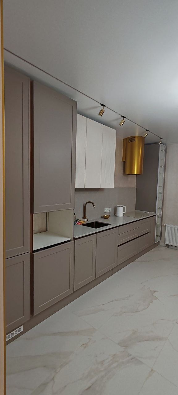 Nowoczesna kuchnia z beżowymi szafkami, złotym okapem i oświetleniem szynowym, widok na umywalkę i blat z akcesoriami.