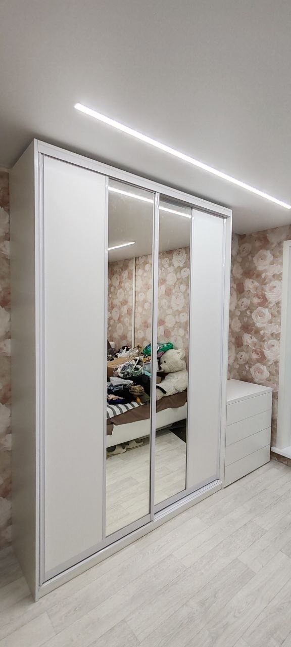 Biała szafa przesuwna z lustrami w sypialni z tapetą w kwiaty, obok komoda z szufladami.