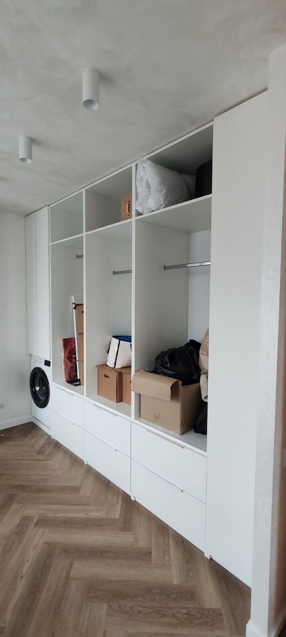 Biała zabudowa meblowa z szufladami i półkami, wkomponowana pralka, na półkach kartony i torby, parkiet w jodełkę na podłodze.