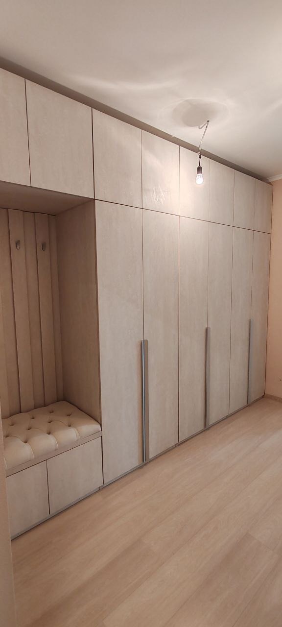 Duża, beżowa szafa wnękowa z siedziskiem i wieszakami, oświetlona wiszącą żarówką bez klosza.