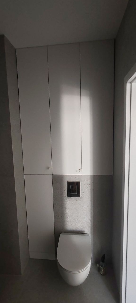 Nowoczesna toaleta z podwieszaną muszlą, zabudowana szafkami w kolorze białym i szarymi płytkami imitującymi beton, z widocznym chromowanym przyciskiem spłukującym.